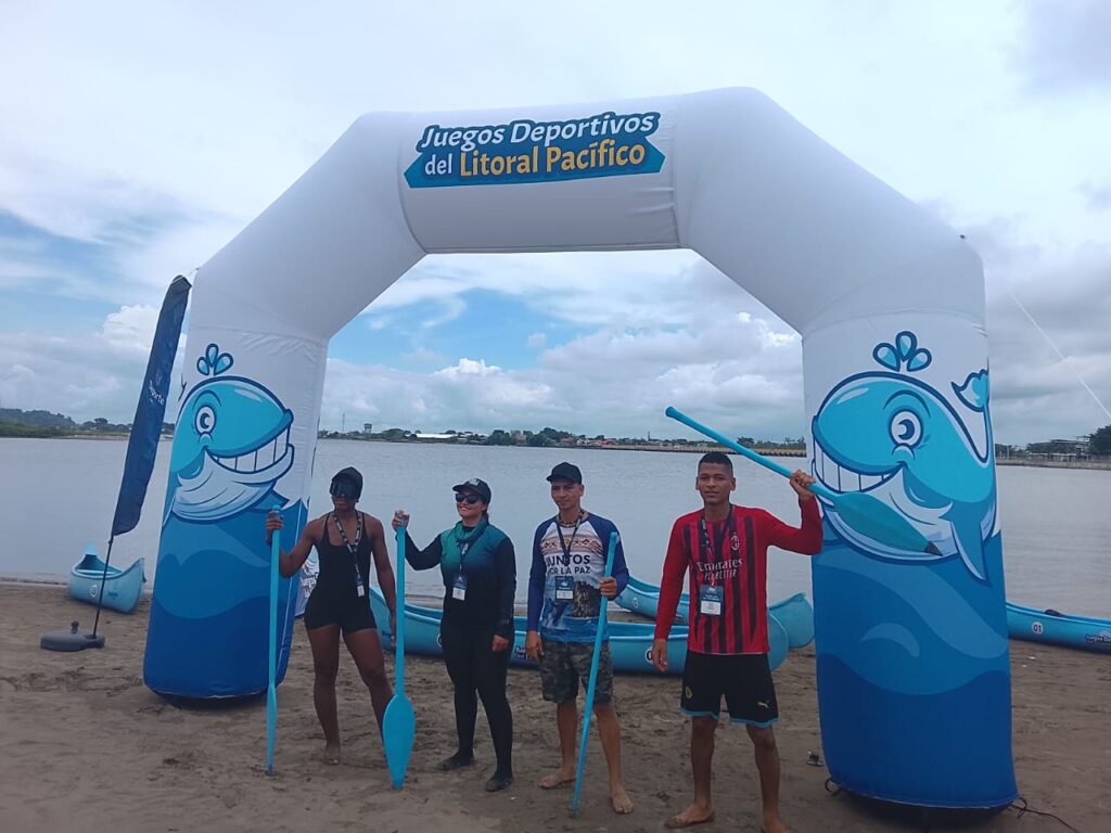Cuatro participantes posan con remos frente al arco oficial de los Juegos Deportivos del Litoral Pacífico, en una zona costera de Tumaco.