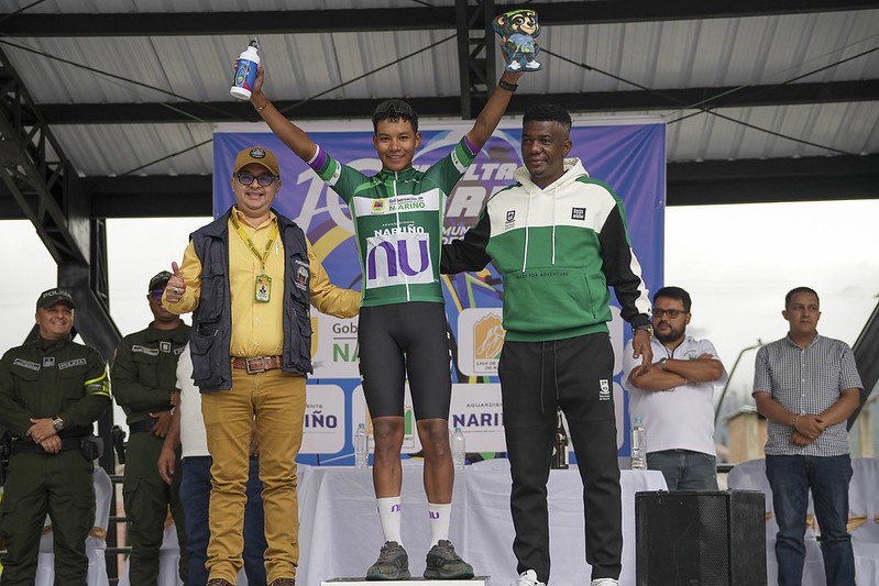 El ciclista Richard Huera en el podio luciendo la camiseta verde de líder de la Décima Vuelta a Nariño, flanqueado por autoridades deportivas y sosteniendo sus trofeos.   