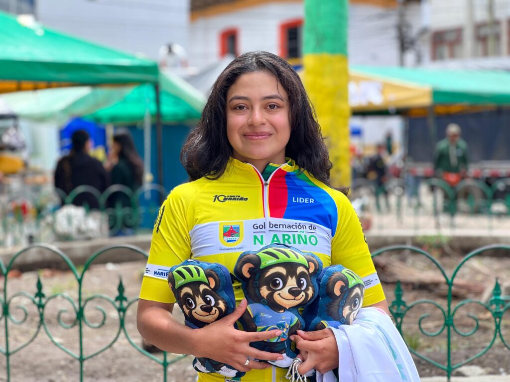 Retrato en plano medio de la ciclista nariñense Anahí Rosero luciendo la camiseta amarilla de líder de la Décima Vuelta a Nariño y sosteniendo tres peluches de la mascota oficial en Iles.