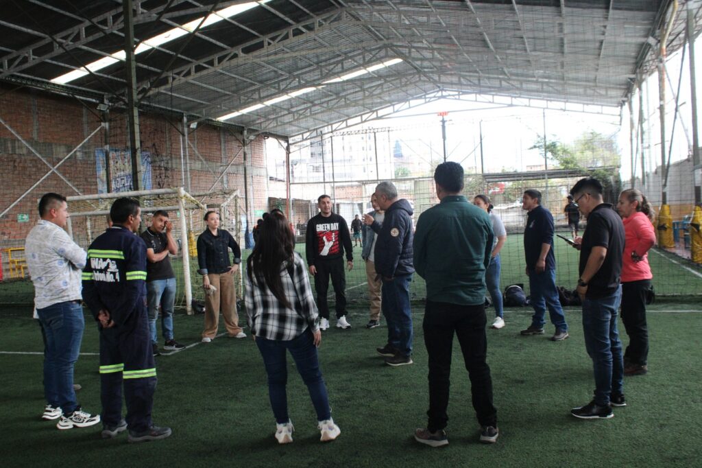 Grupo de participantes en una capacitación práctica parados en círculo sobre una cancha de césped sintético bajo un tinglado cubierto.